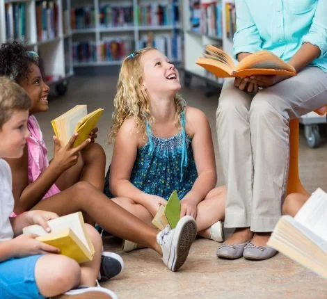 Children sit in a classroom as a teacher reads to them.